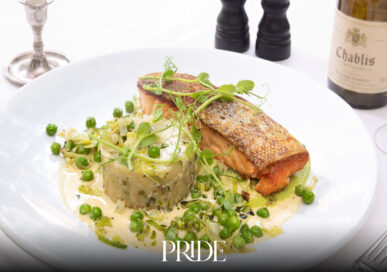 Pan-fried sea trout with crushed new potatoes, peas and leeks on a white plate, with salt and pepper mills and a bottle of Chablis behind